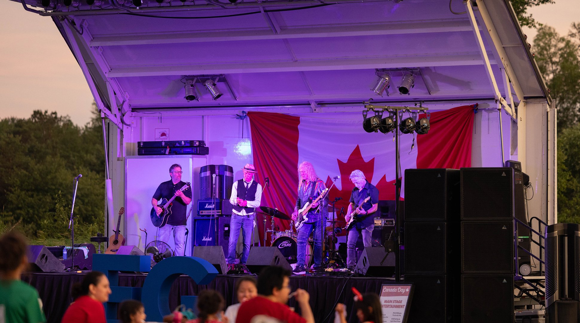 A live band performs on an outdoor stage illuminated with purple lighting, with a large Canadian flag hanging behind the musicians. Several performers play guitars and stand among amplifiers, speakers, microphones, and a drum kit. Audience members are visible in the foreground, and a sign near the stage references Canada Day entertainment. Trees and evening sky surround the venue.