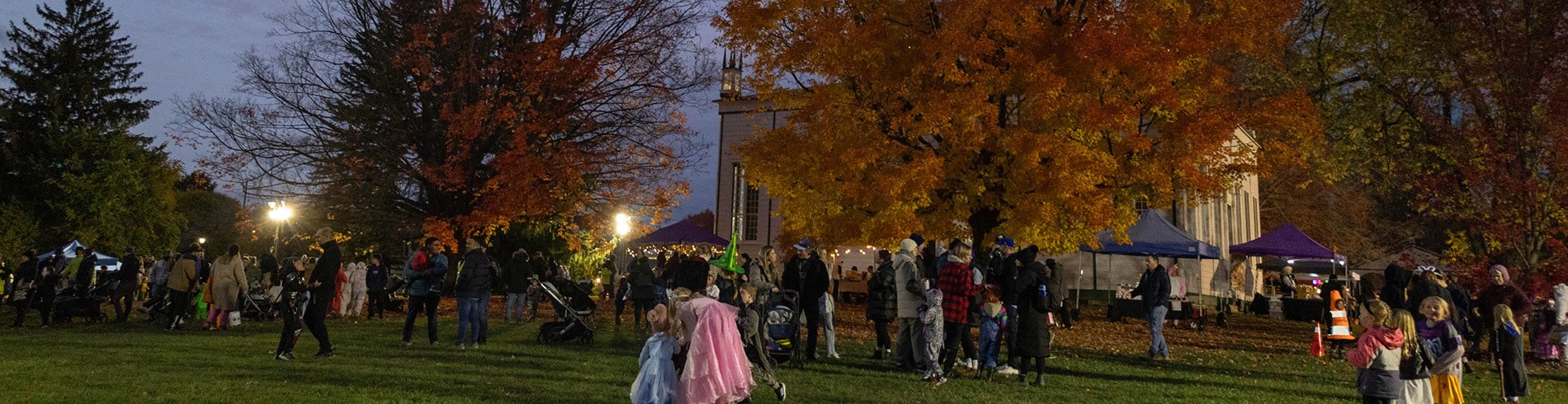 A Halloween-themed community event takes place on a lawn at dusk, with families and children in costumes gathered among large trees displaying vibrant orange and red autumn foliage. Tents and vendor booths are set up near a brightly lit building in the background, while event lights illuminate the crowd and the fall landscape.