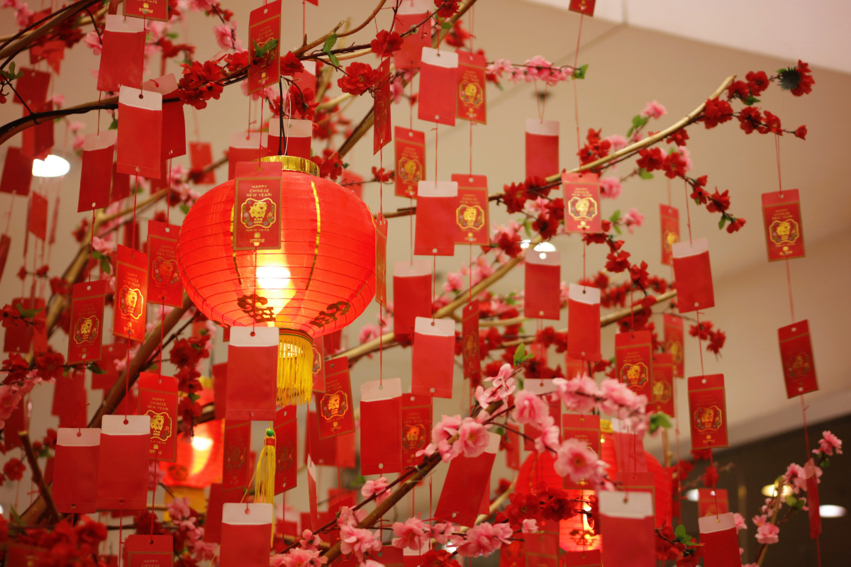 Red lanterns and hanging red envelopes decorate flowering branches with pink and red blossoms in a festive Lunar New Year display. A glowing red lantern is featured in the foreground, surrounded by dozens of suspended red envelopes, creating a vibrant and colourful indoor scene.