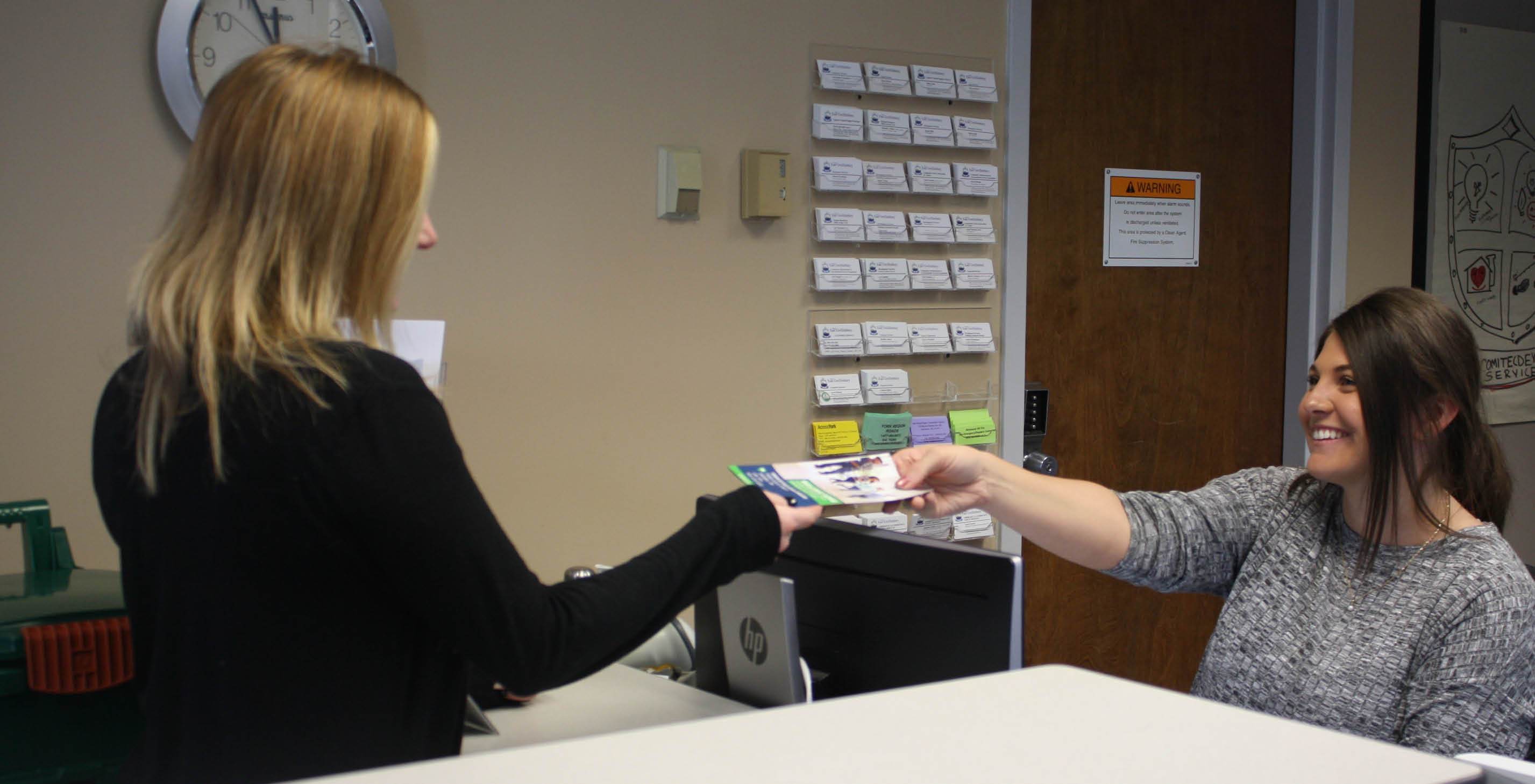 Woman sitting behind counter, handing a brochure to a woman