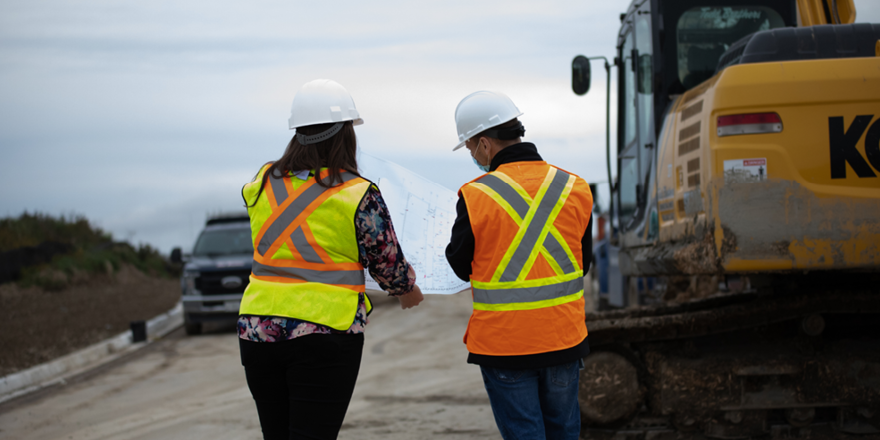 Two construction workers wearing high-visibility safety vests and hard hats review site plans beside a large excavator on a road construction site under an overcast sky.