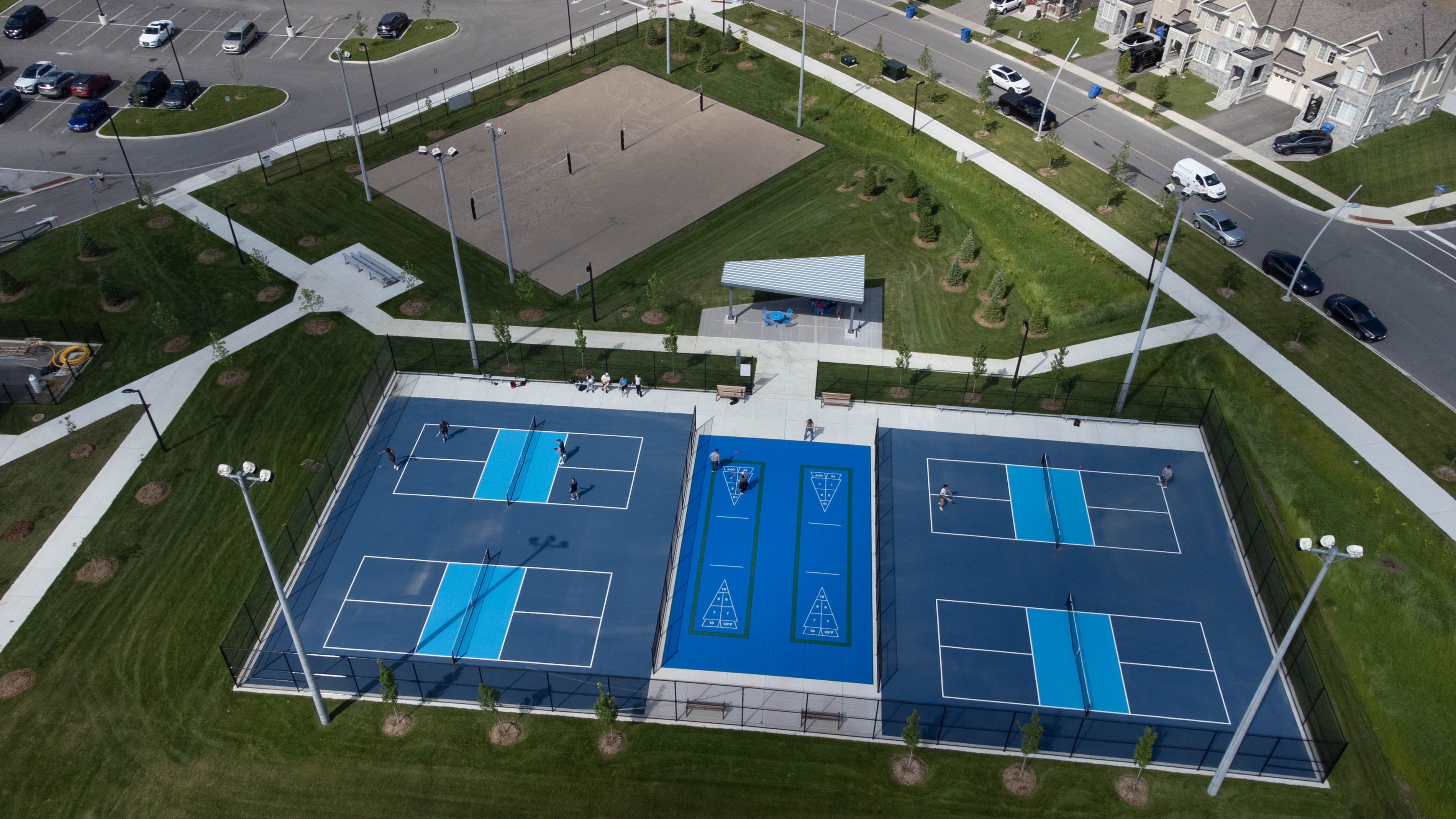 Aerial view of a community recreation area featuring four blue pickleball courts and two blue shuffleboard courts enclosed by black fencing. Several people are playing on the courts. Behind the courts is a sand volleyball court, a covered pavilion with tables, paved walking paths, landscaped grass slopes, and young trees. Residential homes and parked vehicles line the surrounding streets.  Provide your feedback on BizChat