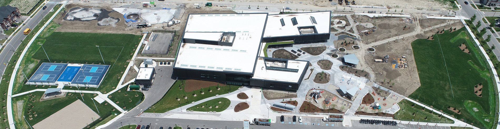 Aerial panoramic view the Health and Active Living Plaza. A large modern building with white roofs sits at the center, surrounded by landscaped walkways, parking areas, playgrounds, sports fields, and four blue outdoor pickleball/tennis courts. Construction and landscaping work are still underway in several areas, with bare soil, equipment, and newly planted trees visible around the site. Residential streets and homes border the complex.