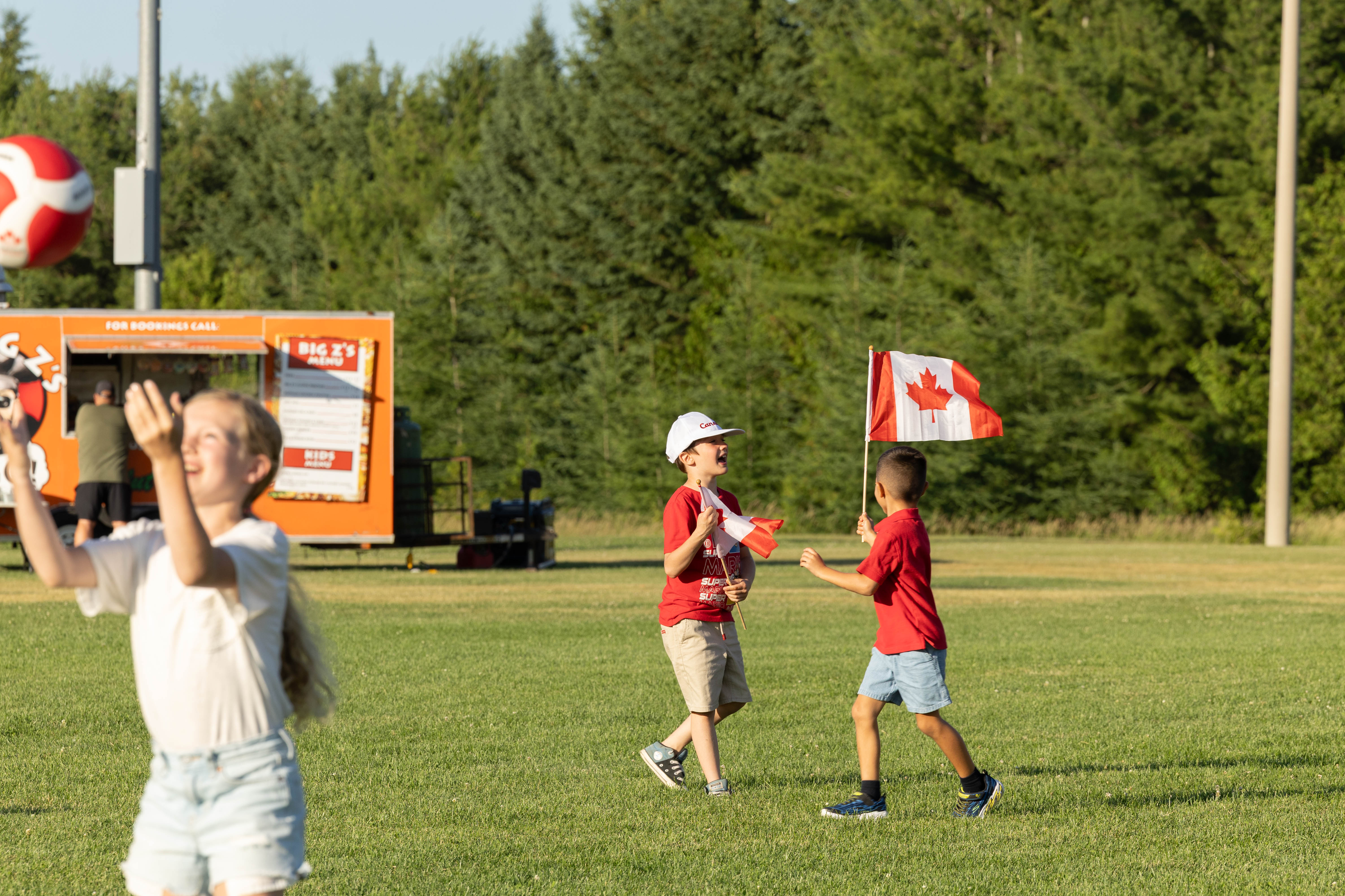 Several people enjoy activities on a grassy field during a community celebration. In the foreground, a person in a white shirt reaches up toward a red-and-white beach ball. In the background, two children wearing red shirts play while holding small Canadian flags, with one child also carrying a larger Canadian flag. An orange food truck and a line of green trees are visible behind them in the late-afternoon sunlight.