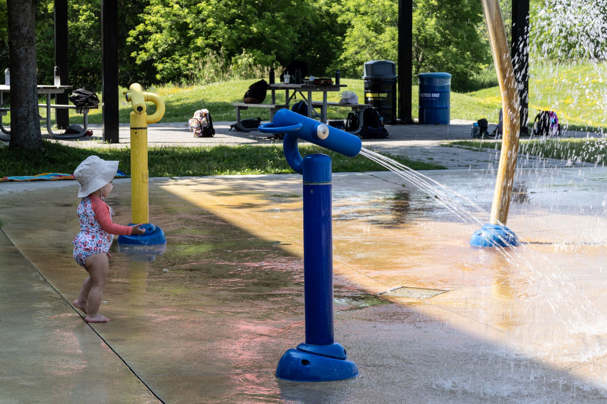 A young child stands barefoot on a splash pad beside blue and yellow water play features spraying streams of water. The splash pad is located in a park setting with a covered picnic area, tables, and green trees visible in the background on a sunny summer day.