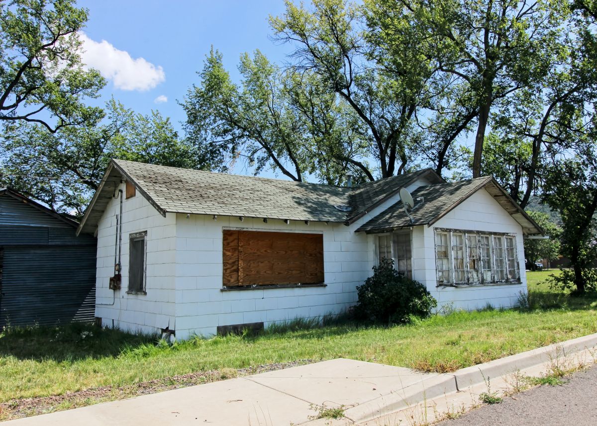 A small, white, single-story house sits on a grassy lot beside a sidewalk. Several windows and the front opening are covered with plywood, and the house appears vacant and neglected. Mature trees tower over the property, casting light shade under a clear blue sky. A corrugated metal building is partially visible on the left side.