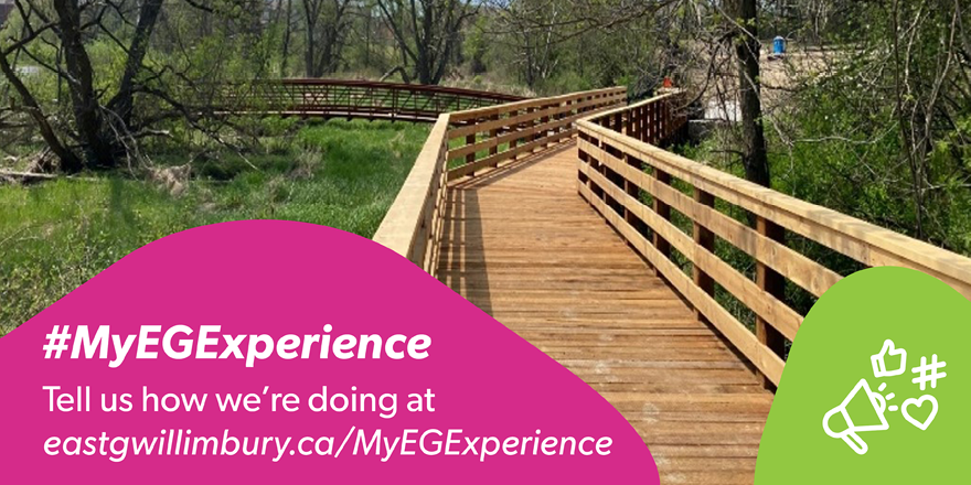 Wooden boardwalk with railings winding through a green wooded wetland area on a sunny day. A pink banner overlays the bottom left of the image with the text “#MyEGExperience” and “Tell us how we’re doing at eastgwillimbury.ca/MyEGExperience.” A green circle on the bottom right contains white icons of a megaphone, thumbs-up, and heart.