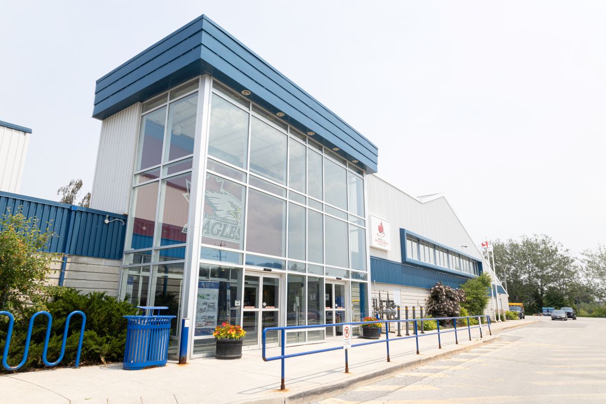 Exterior view of the East Gwillimbury Sports Complex, featuring a large glass-front entrance with blue and white exterior panels, accessible ramp and railings, planters with flowers, bicycle racks, and a parking area along the side of the building.