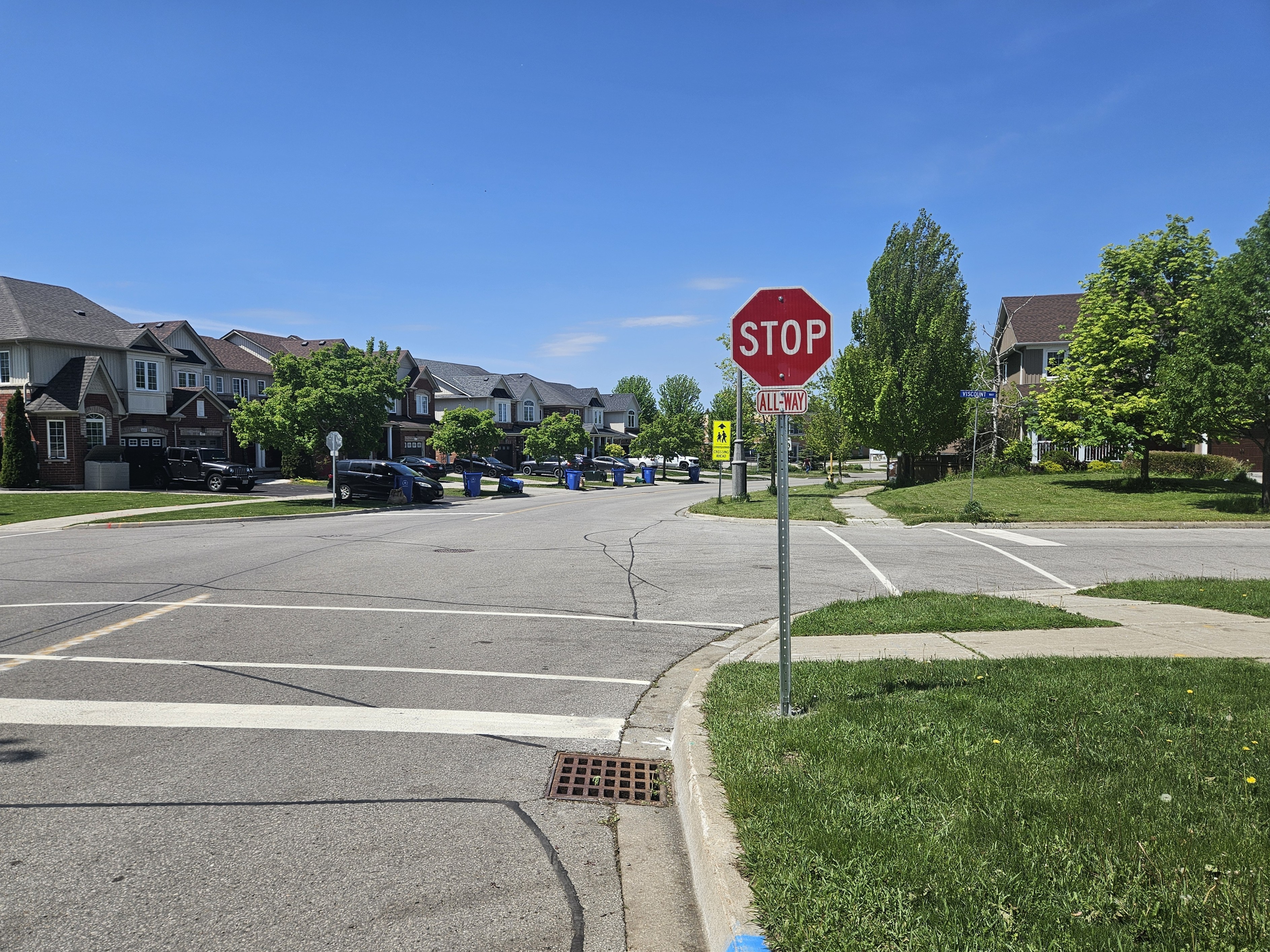 Four-way stop intersection in a residential neighbourhood with a red stop sign and all-way plaque in the foreground. Sidewalks, crosswalk markings, detached homes, parked vehicles, blue recycling bins, and green trees line the streets under a clear blue sky.