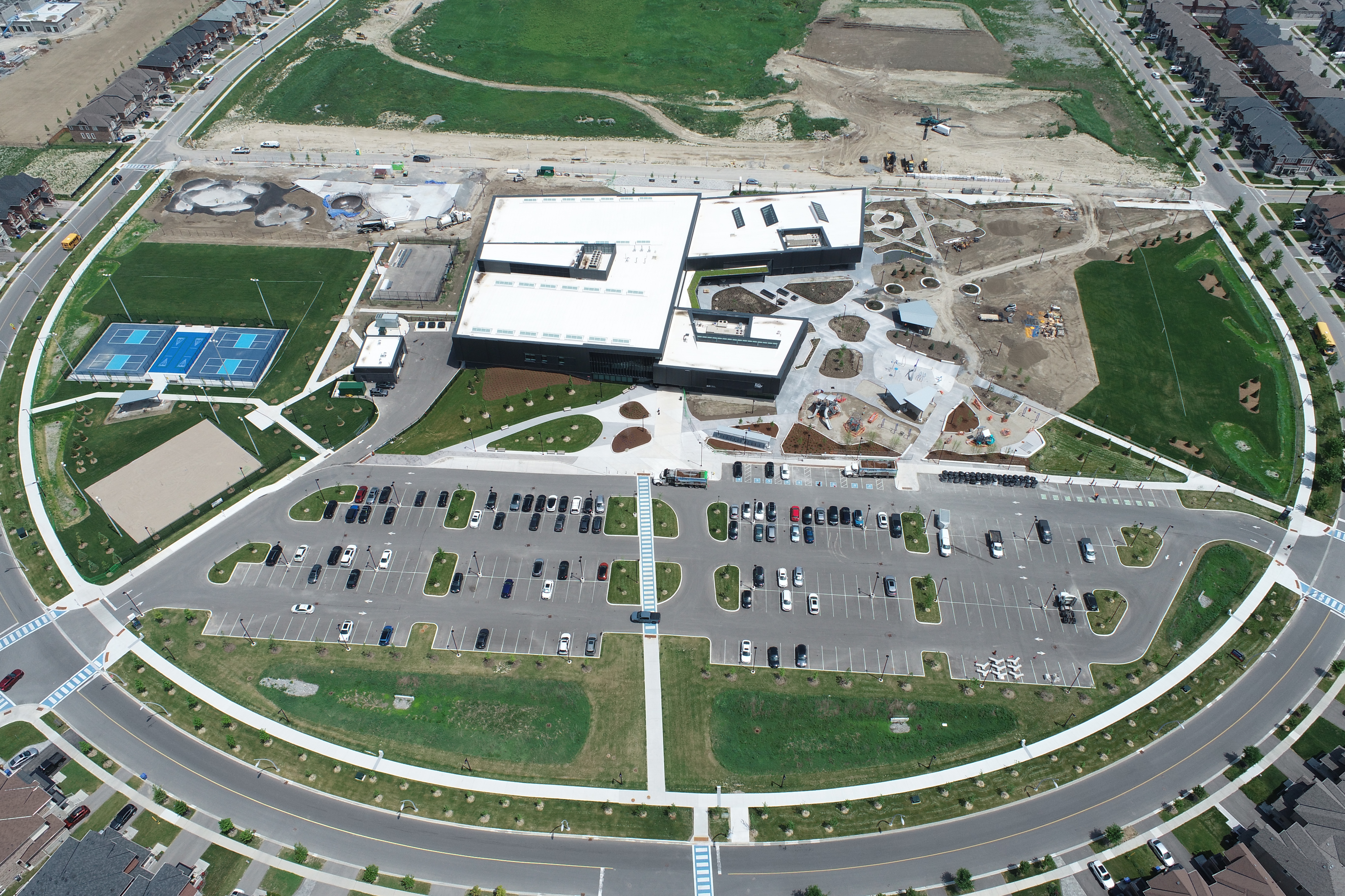 A high-resolution aerial drone view of a large modern community recreation complex surrounded by suburban development. The building has a white roof with several skylights and dark exterior walls. In front of the facility is a large parking lot with numerous parked cars and landscaped islands. To the left are two blue tennis or pickleball courts and a rectangular sand court. To the right of the building is an outdoor plaza and playground area with winding concrete paths, circular planting beds, and a small pavilion. Sports fields with tall light poles are visible on both sides of the complex. Roads and sidewalks curve around the site, with rows of newly built houses along the perimeter. Behind the building, active construction areas, earth-moving equipment, and undeveloped grassy land indicate ongoing site development. The image is taken on a clear day with bright sunlight and minimal shadows.
