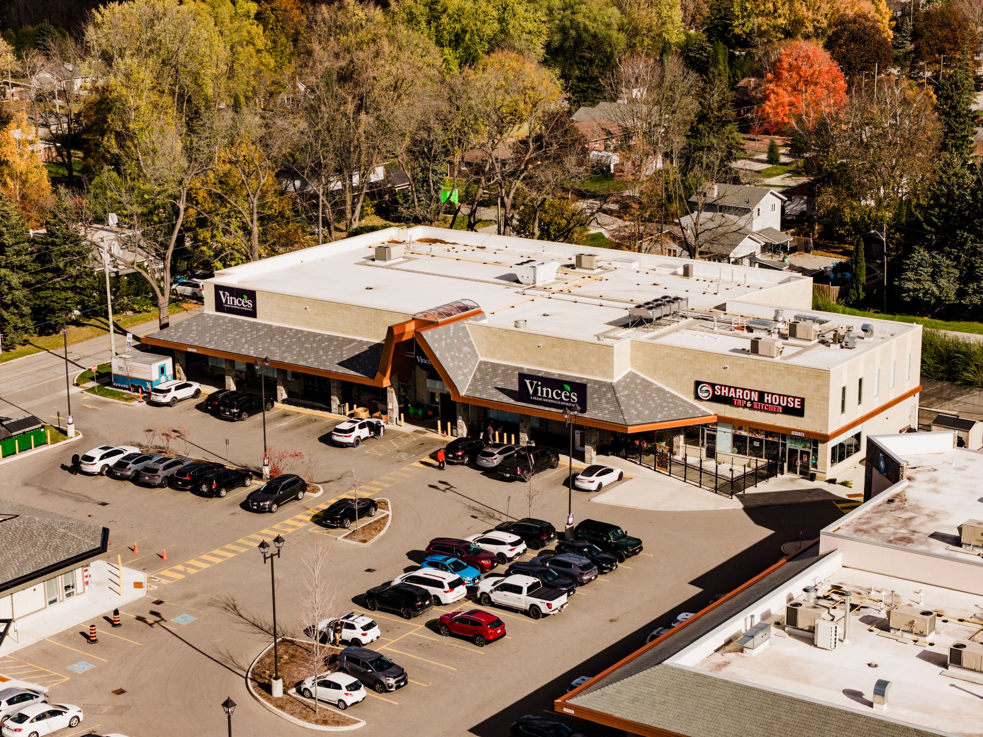 Aerial view of a suburban retail plaza with a large beige commercial building surrounded by a parking lot filled with parked cars. Storefront signs reading “Vince’s” and “Sharon House Thai Kitchen” are visible on the front of the building. Tree-lined residential streets with houses and colorful autumn foliage fill the background. The scene is brightly lit by sunlight, with long shadows cast across the parking area.