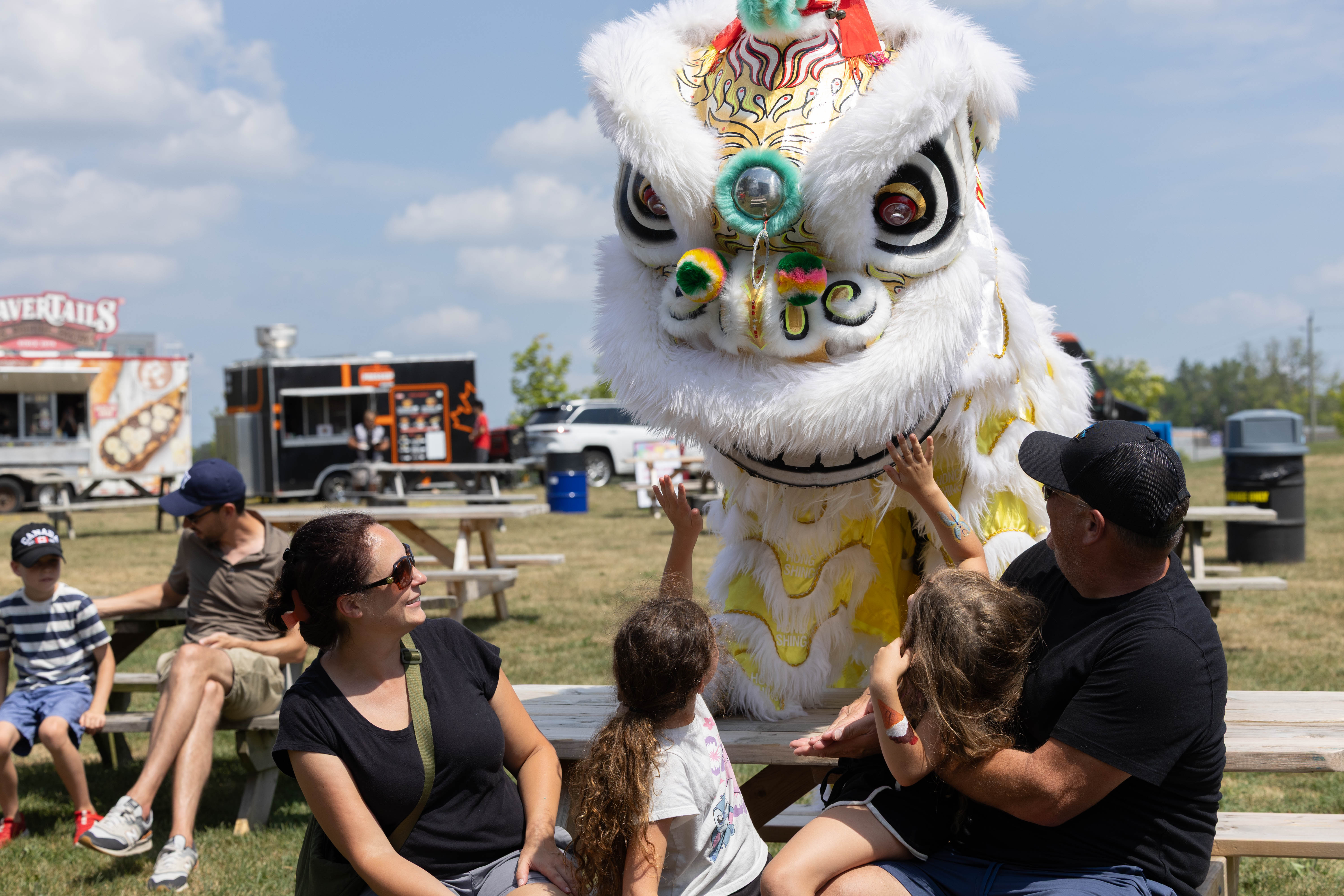 A large white and yellow lion dance costume stands beside a picnic table at an outdoor community event. Several children reach out to touch the lion while adults sit nearby. Food trucks, picnic tables, and open grassy space are visible in the background under a bright, partly cloudy sky. The image captures a cultural performance and family-friendly festival atmosphere