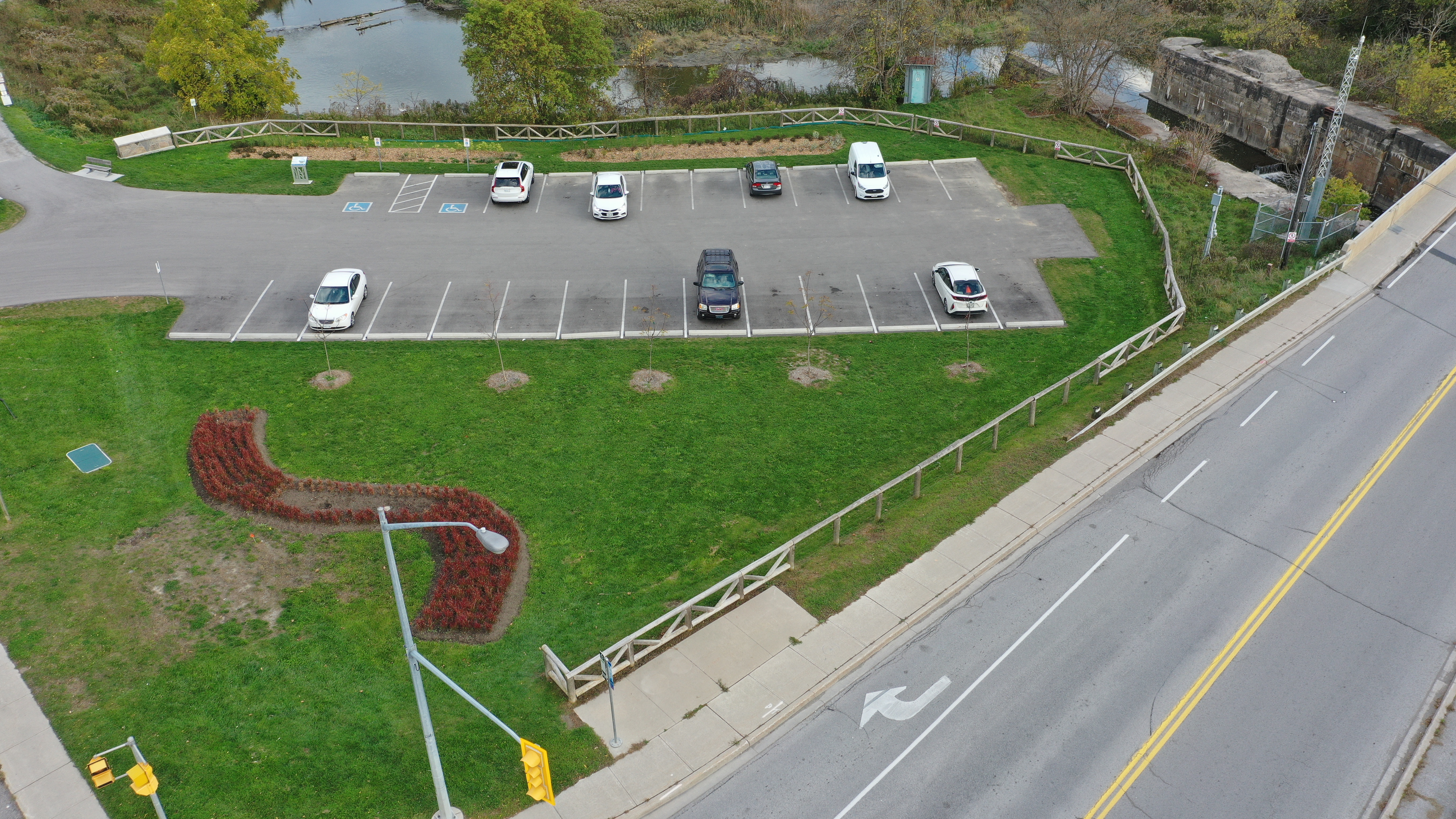 Aerial view of a partially occupied parking lot for the Nokiida trail, beside a wetland and historic lock structure. Several cars and a white van are parked in marked spaces, including accessible parking spots. The lot is bordered by grass, wooden fencing, and landscaped flower beds. A two-lane road with a turning-arrow marking runs along the right side of the image, while trees, water, and marsh vegetation fill the background.  Provide your feedback on BizChat