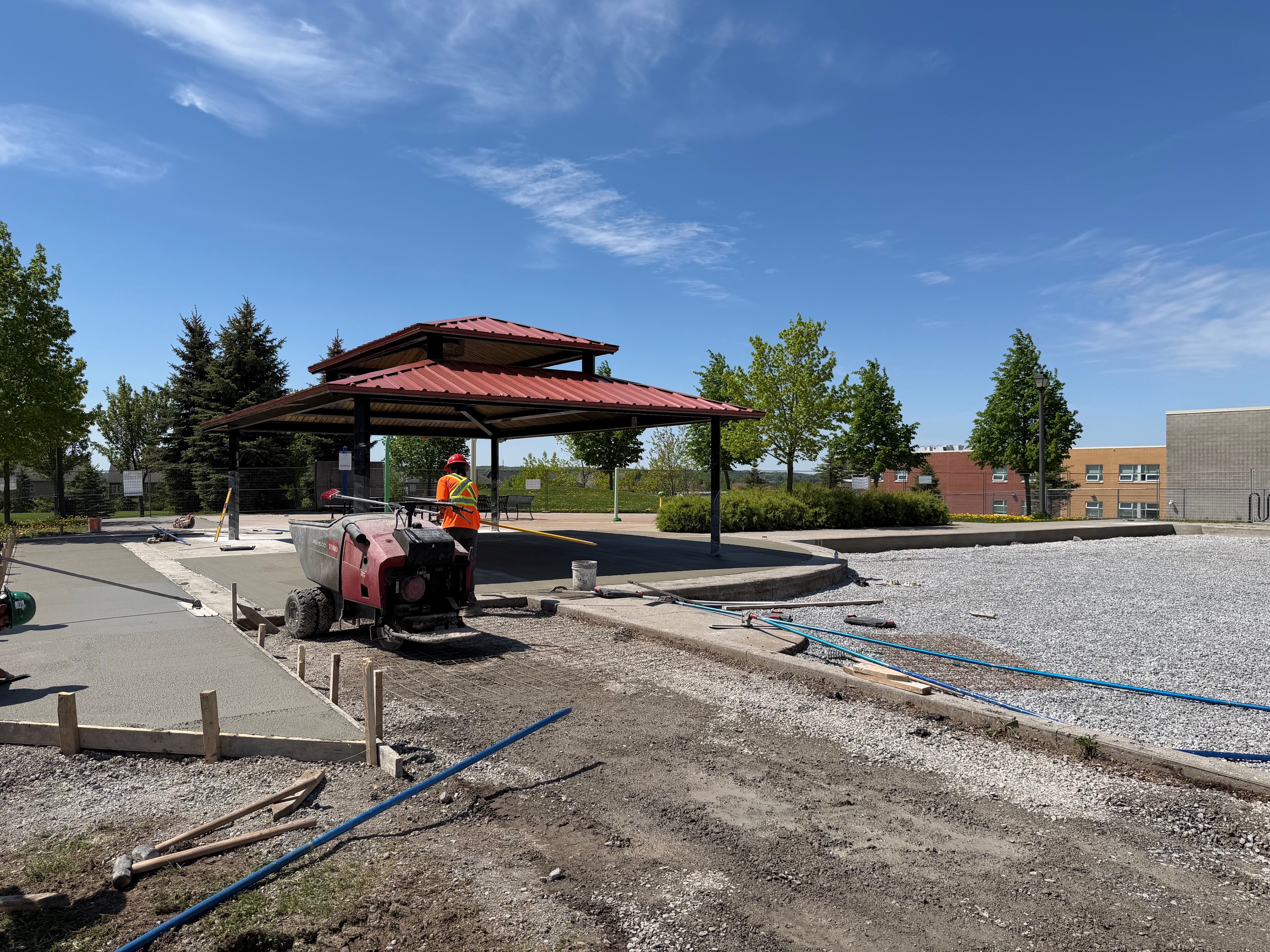 Outdoor construction site with a worker operating a concrete paving machine near a red-roofed pavilion, fresh concrete forms in the foreground, and trees and buildings in the background under a clear blue sky