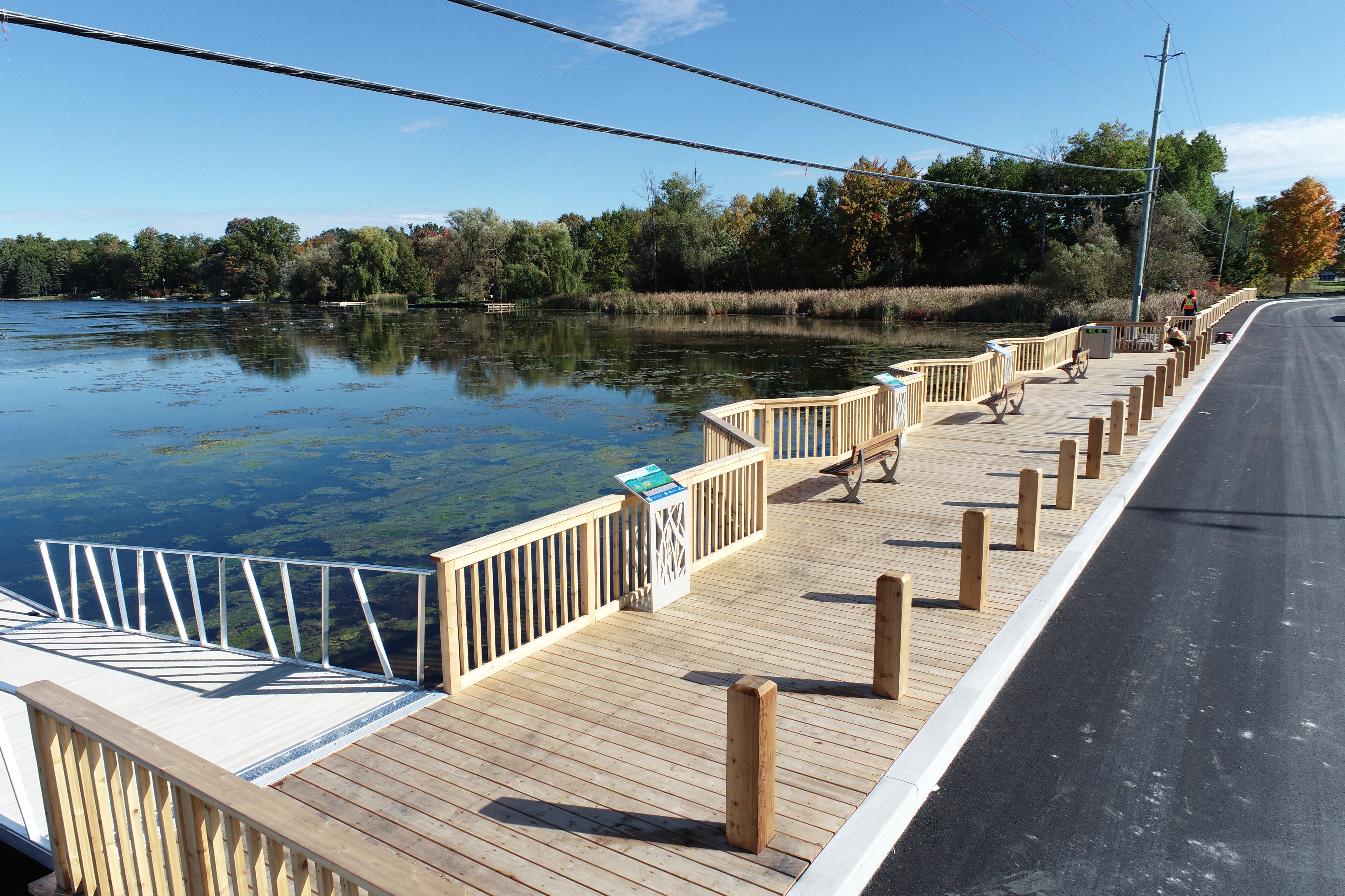 A newly completed wooden waterfront boardwalk and viewing area along the edge of a calm river. The boardwalk features railings, benches, interpretive signage and a dock access point, running parallel to a paved roadway. Naturalized shoreline vegetation and mature trees line the opposite bank under a clear blue sky.